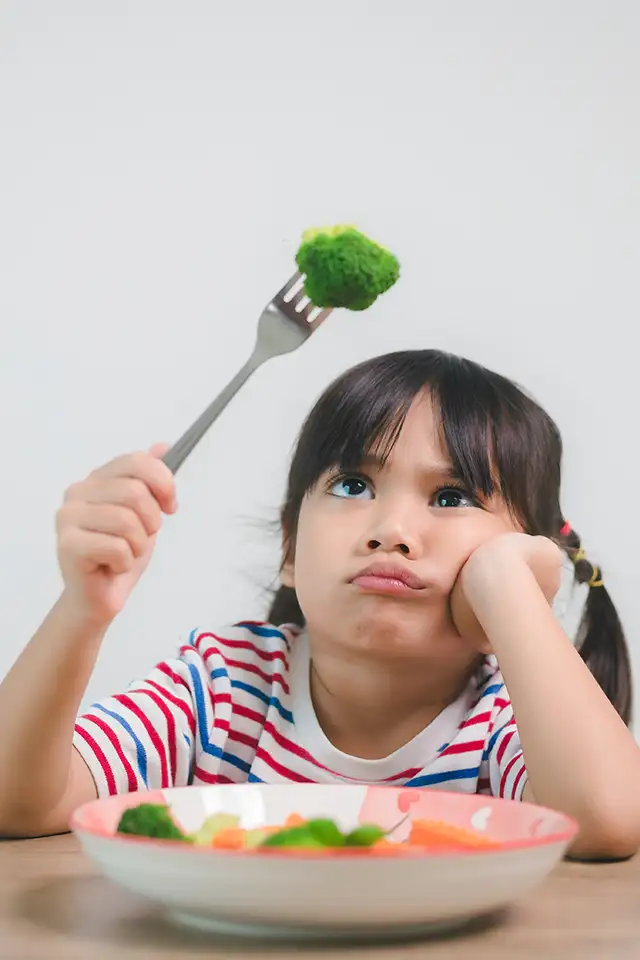 A picky eater, a young girl stares begrudgingly at the piece of broccoli on the end of her fork.
