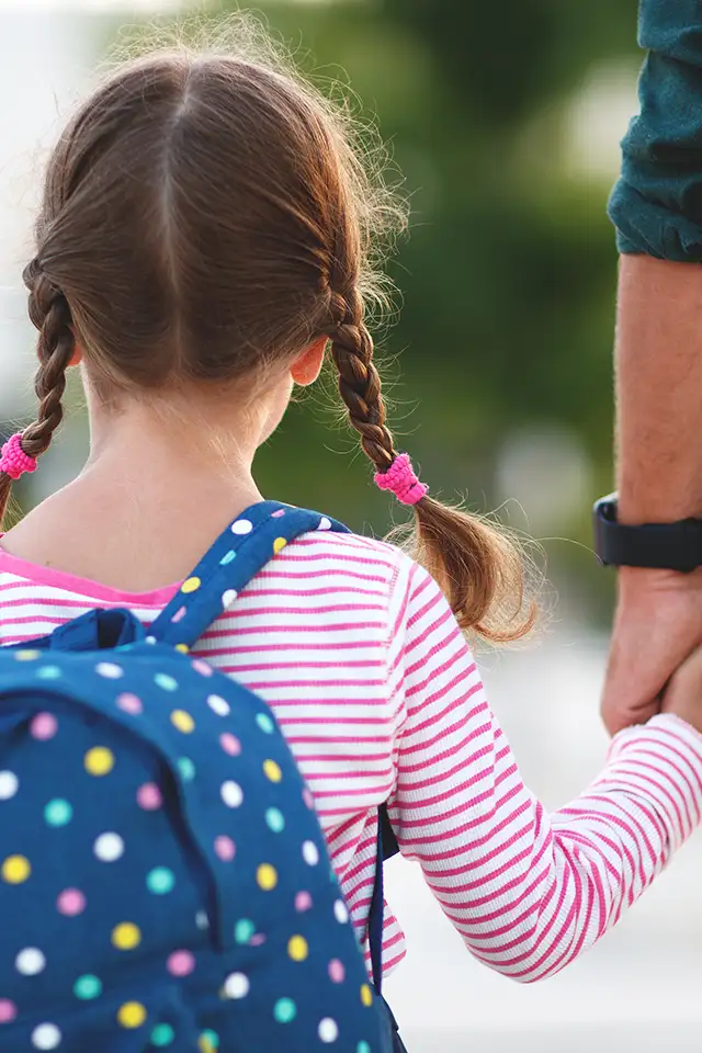 A young girl with a backpack on holds her father's hand as they prepare to walk into a school.